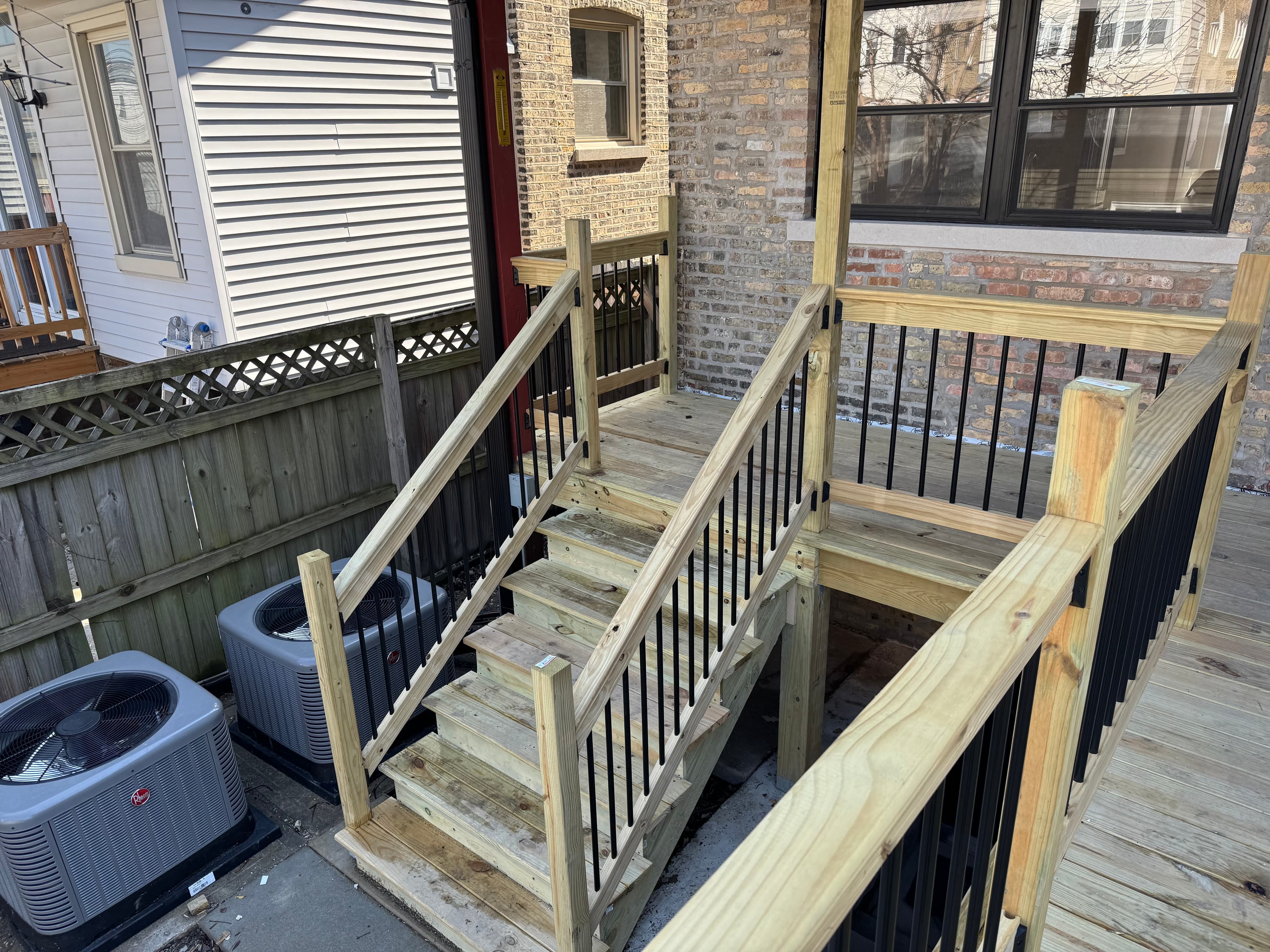 New pressure-treated porch stairs and landing with black metal balusters beside a brick wall