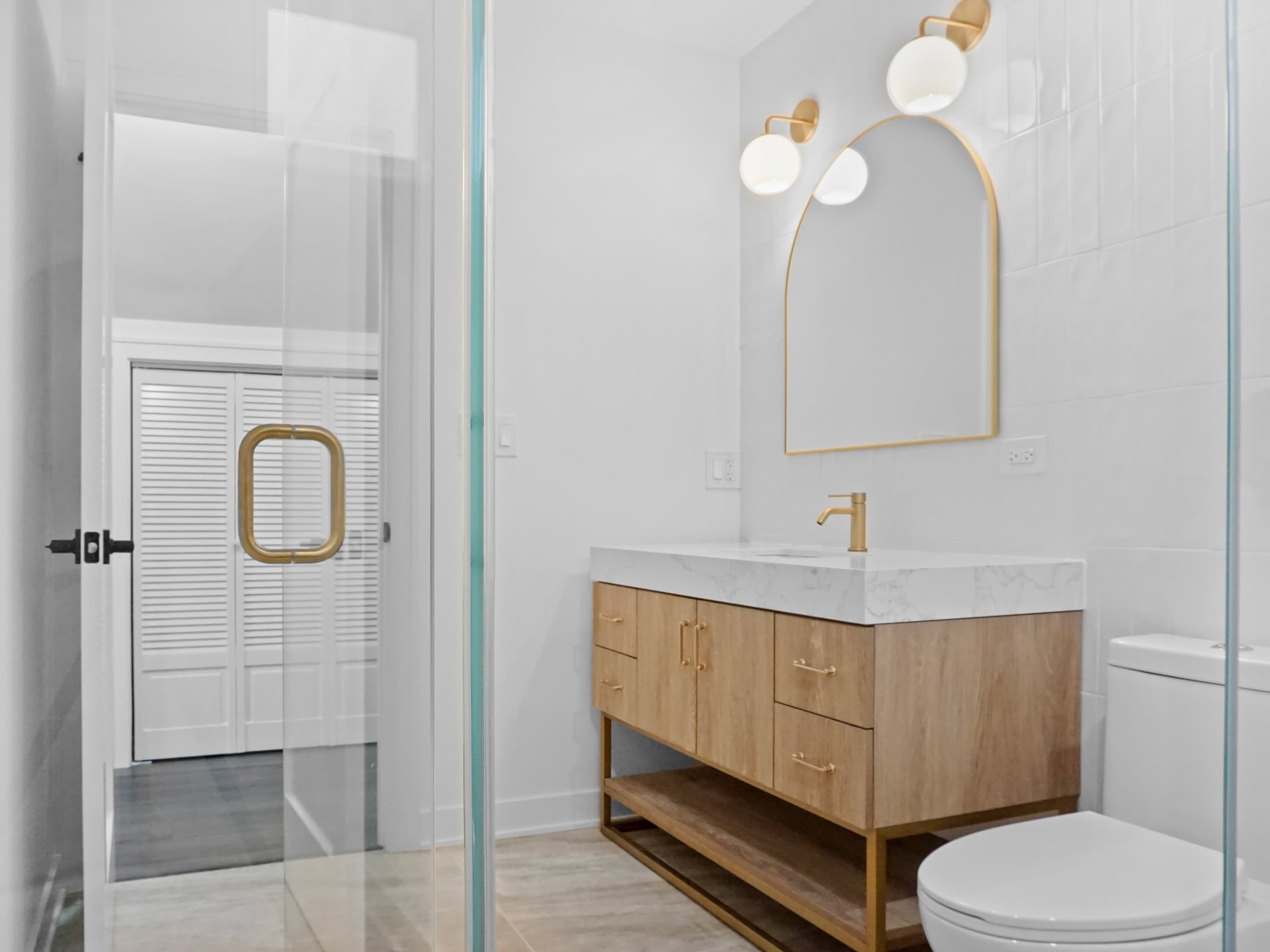 A bathroom with an arched gold mirror, globe sconces, and a floating wood vanity